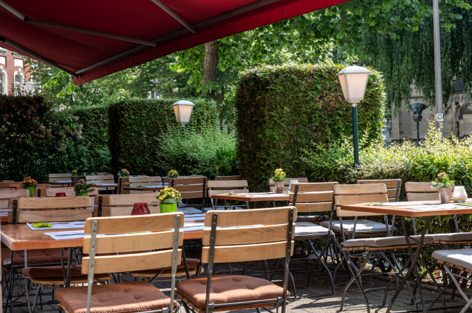 Außenbereich von La Taverna: Tische, grüne Hecke, Bäume und rote Sonnenschutzmarkise